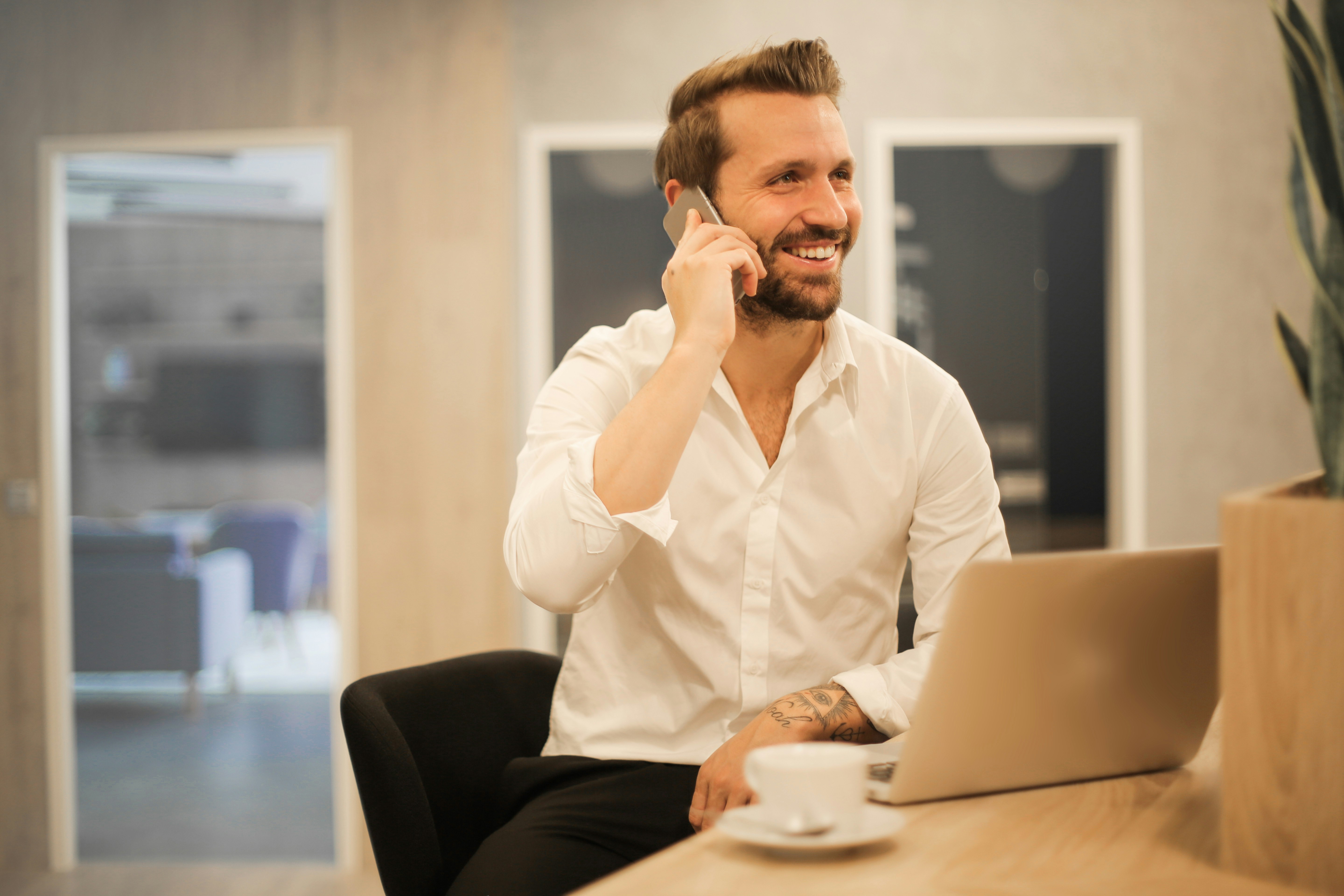 Man in a white shirt smiling while on a phone call at his desk with a laptop