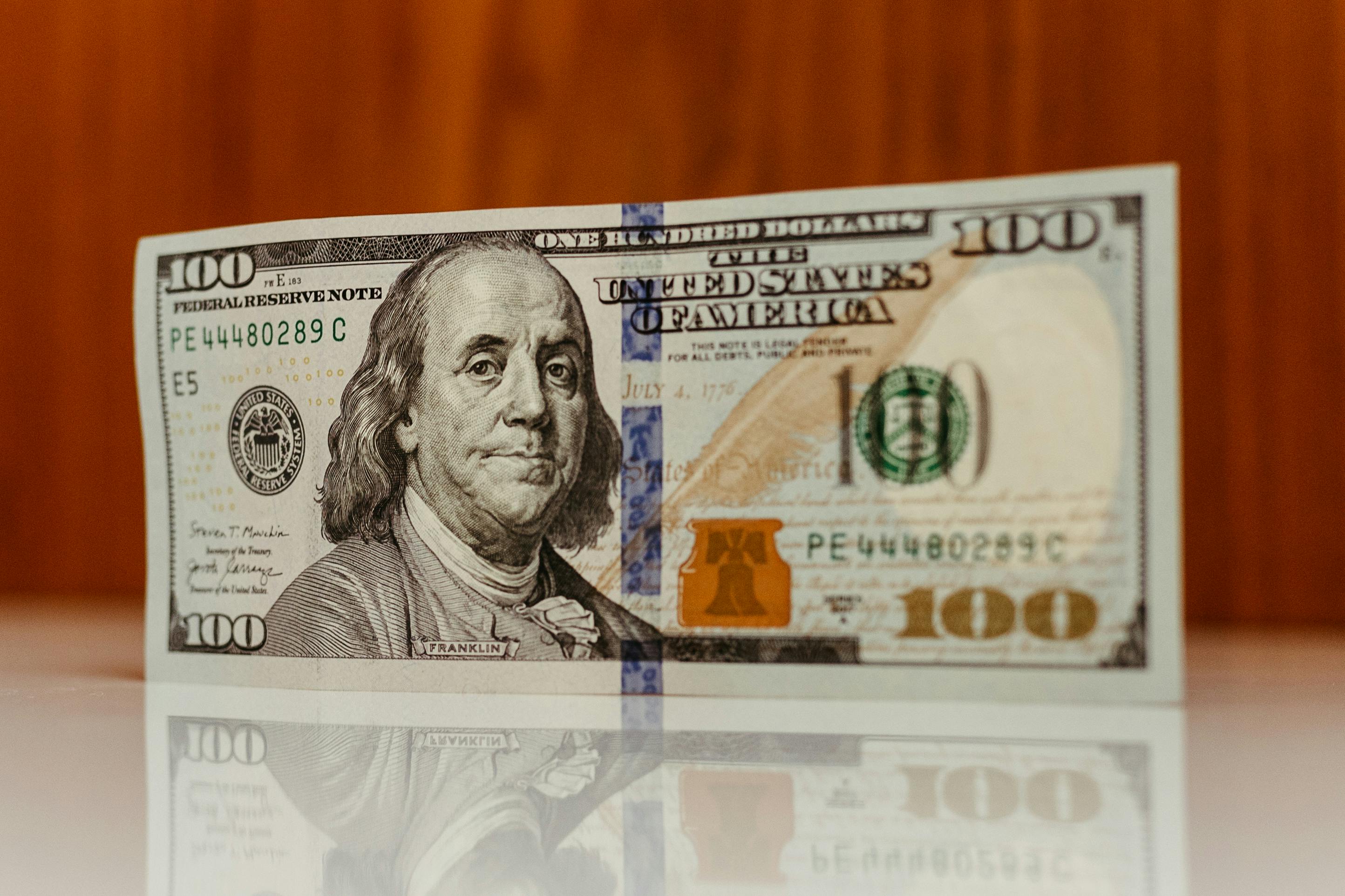 A $100 US bill standing upright on a reflective white surface