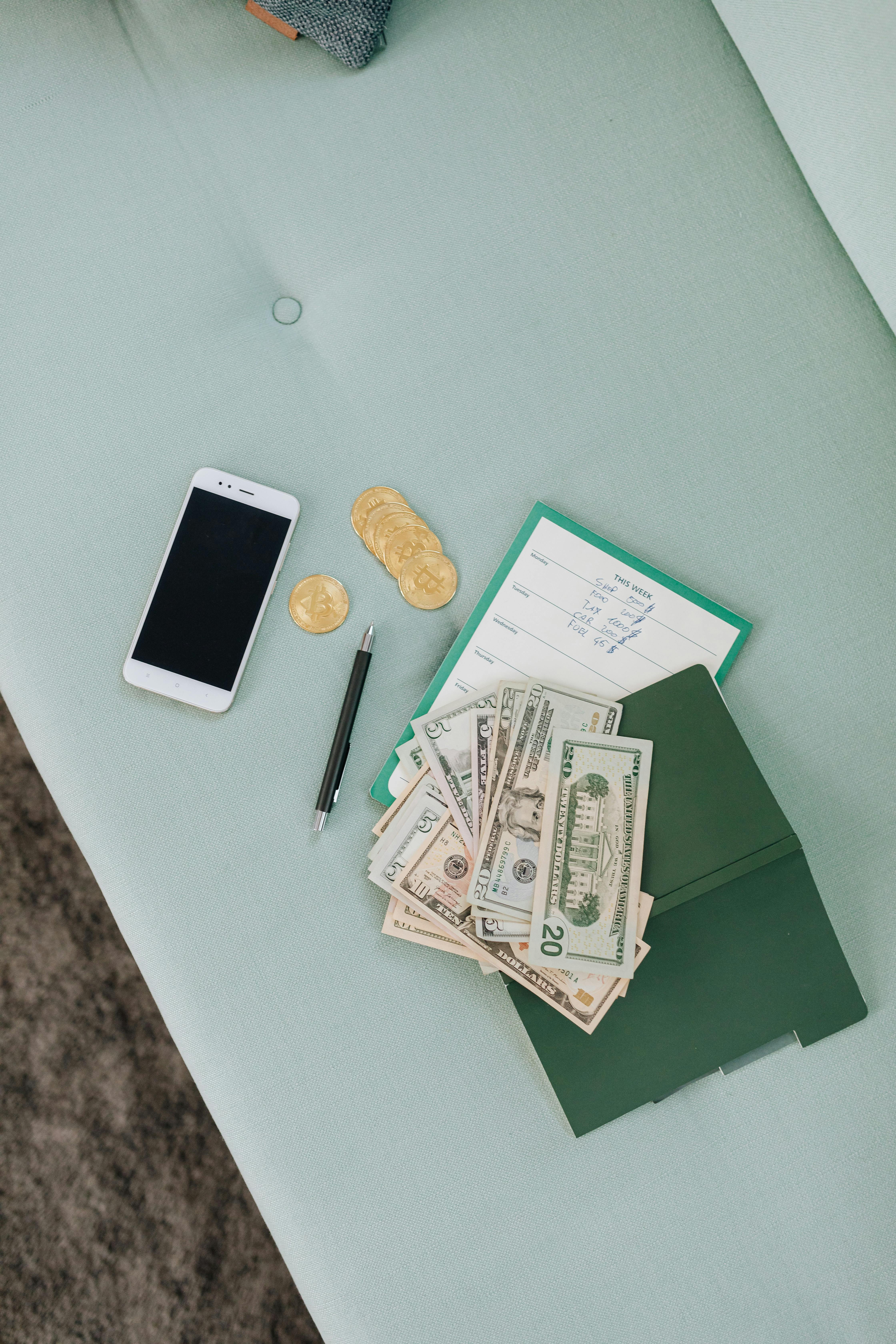 Flat lay of cash, gold coins, a smartphone, and a green checkbook on a light blue surface