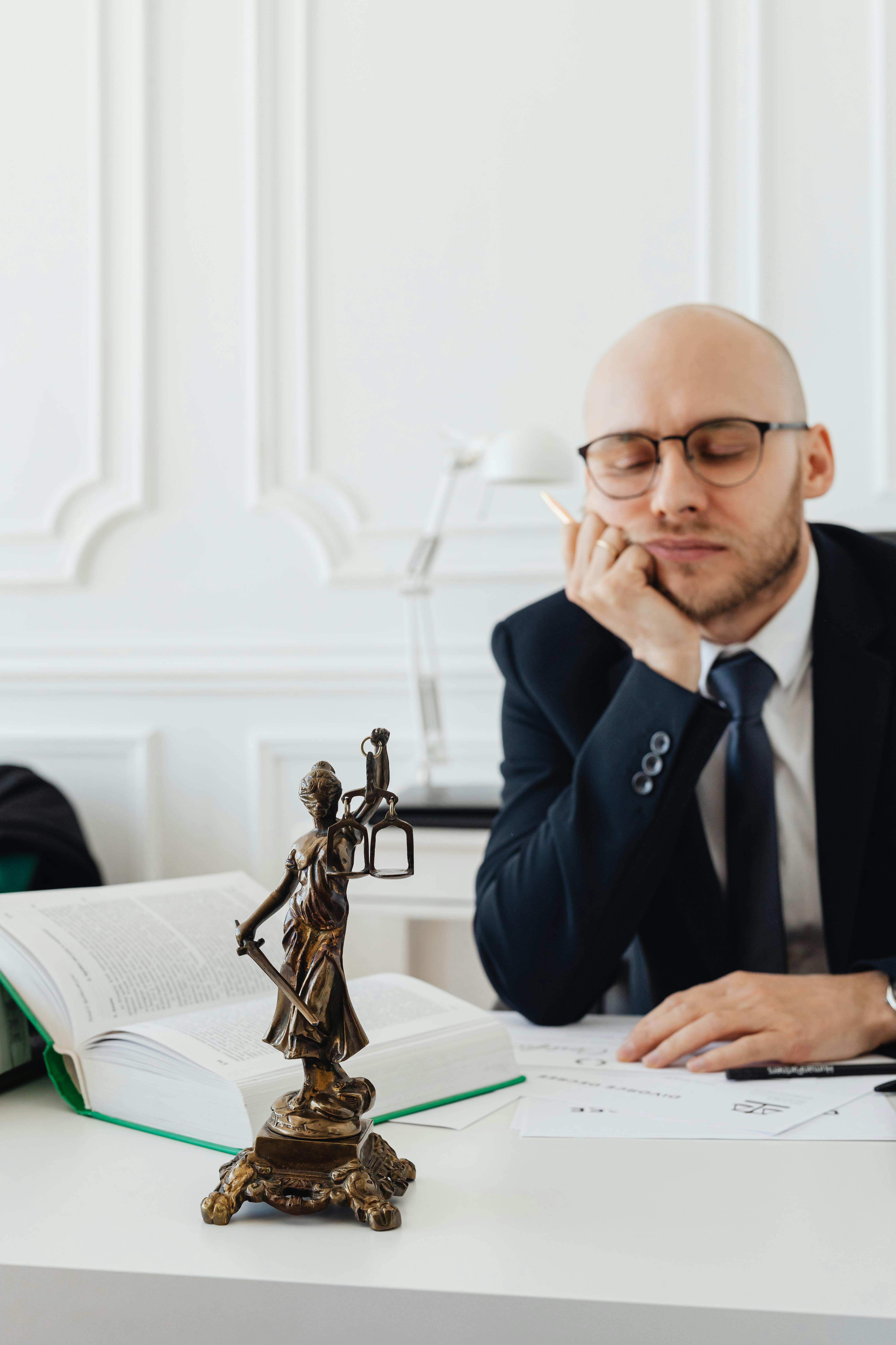 Attorney in a suit and glasses sitting thoughtfully at a desk with a Lady Justice statue and open law book