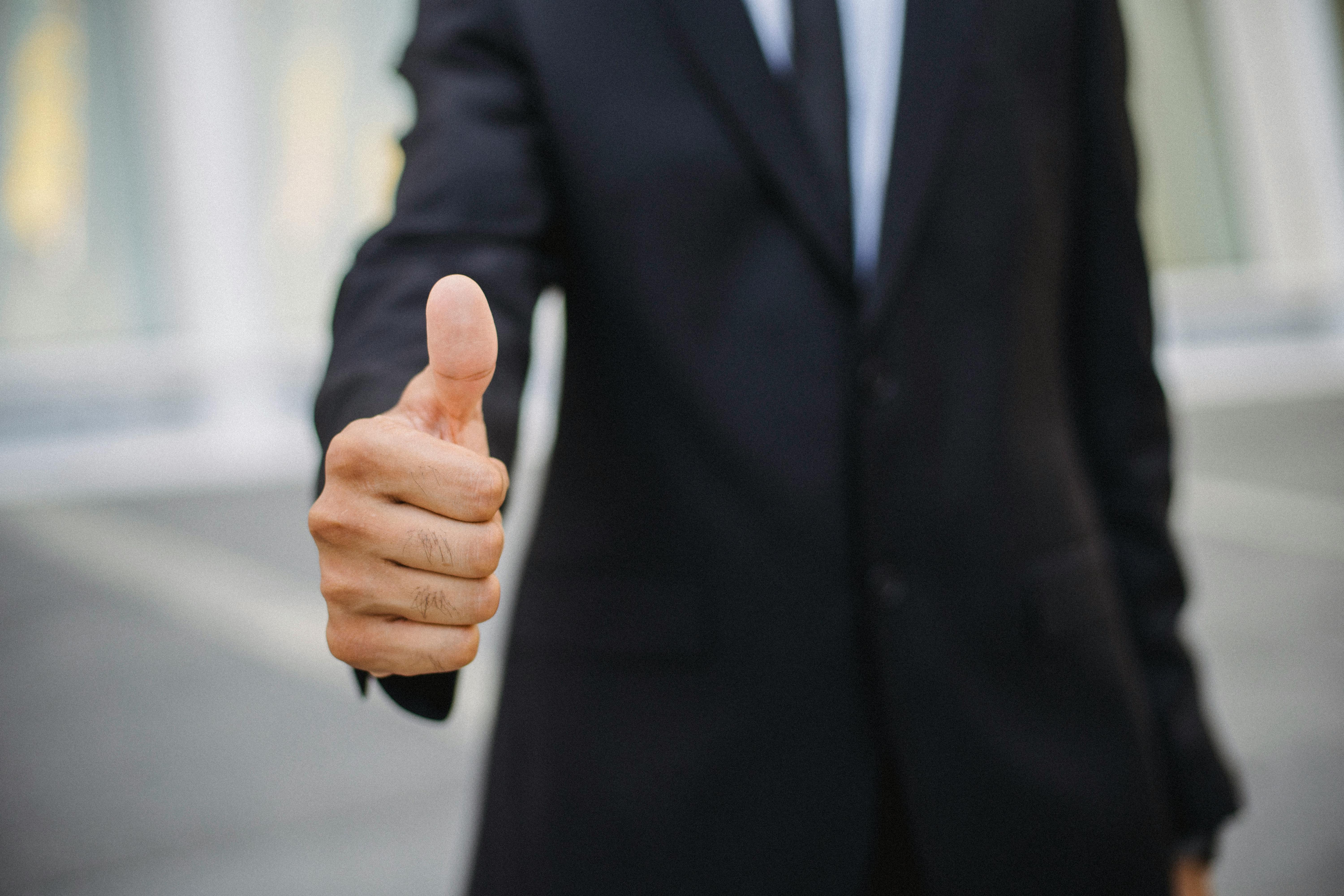Man in a dark suit giving a thumbs up gesture toward the camera outdoors