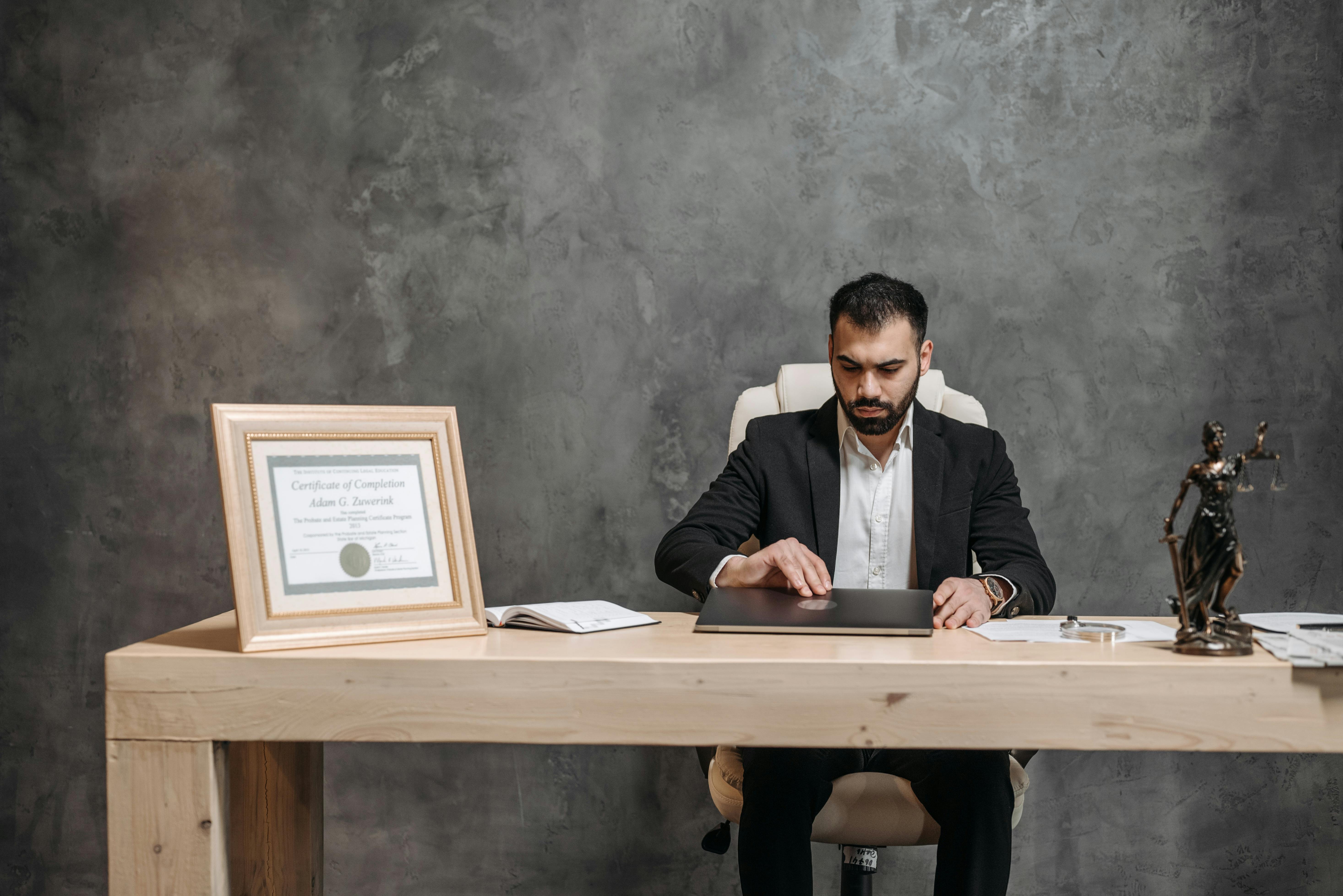 Attorney working at a desk with a laptop, framed certificate, and a Lady Justice statue