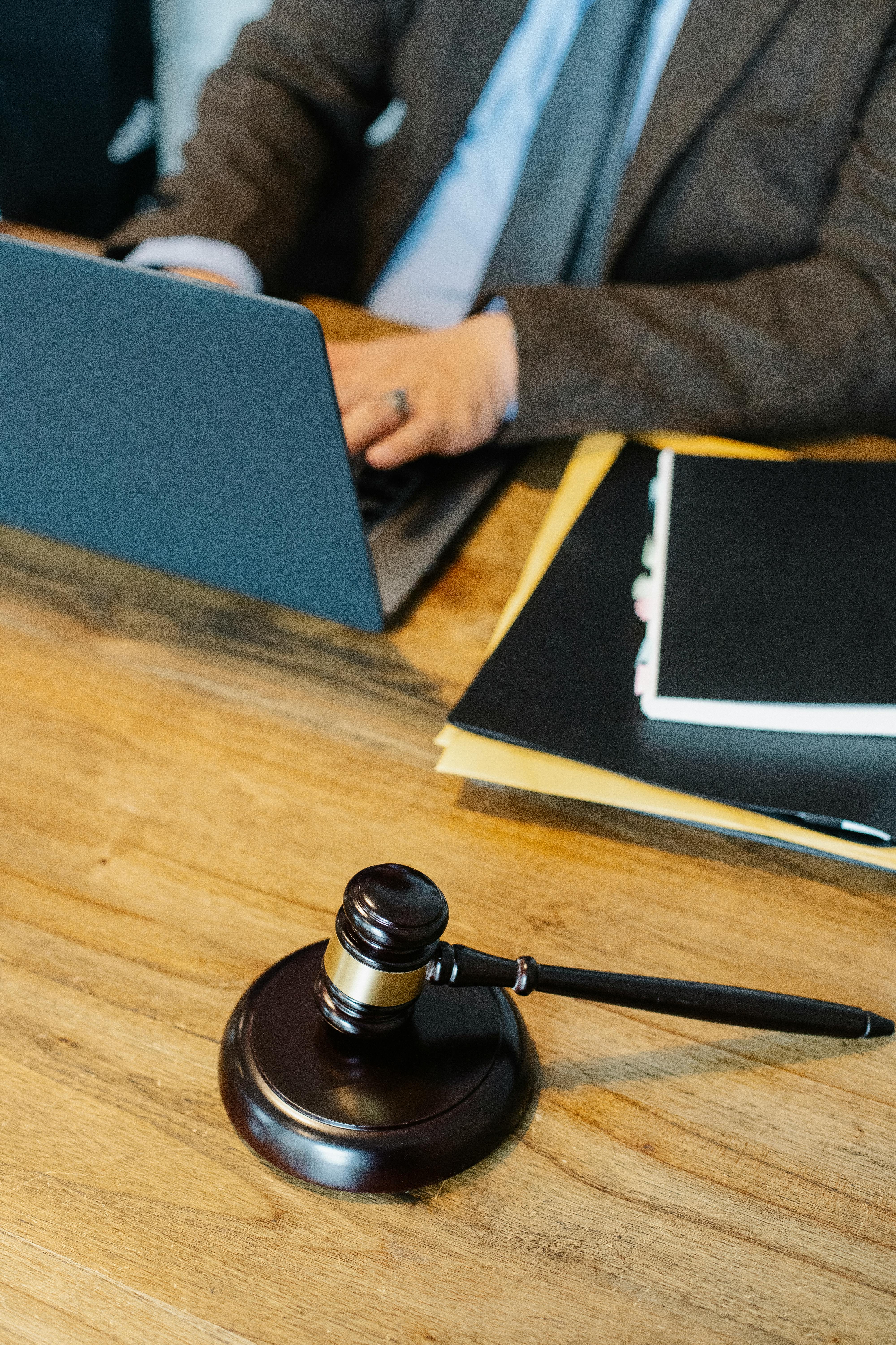 Judge's gavel in the foreground on a wooden desk with an attorney working on a laptop in the background