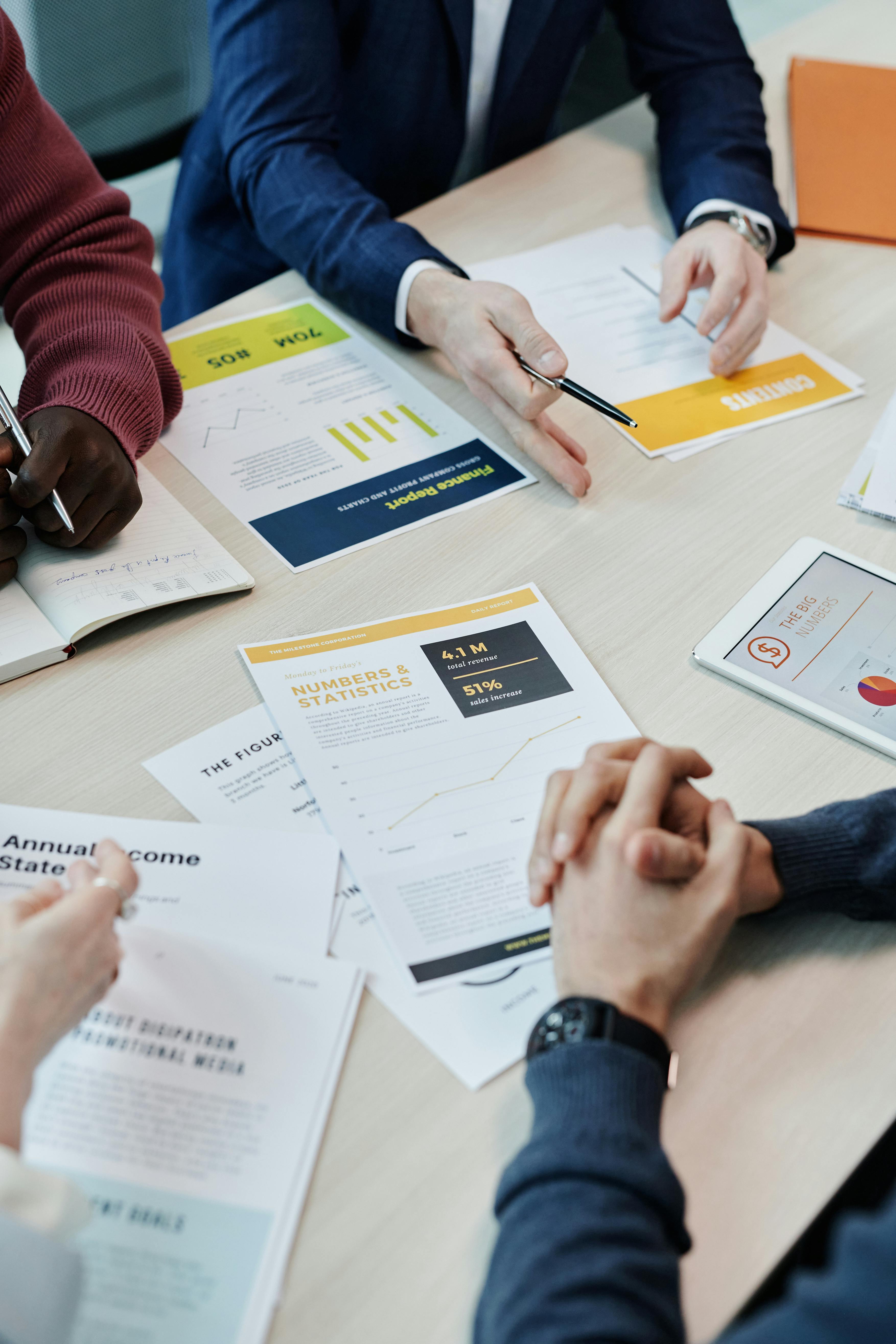 Multiple people around a conference table reviewing printed finance reports and statistics documents